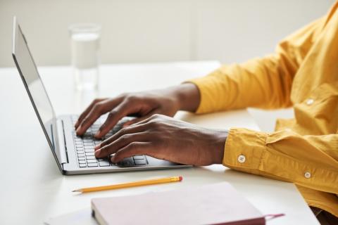 A man's hands, with a glimpse of yellow shirt, typing on the keyboard of a laptop on top of a white table.