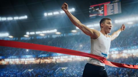 A marathon runner crosses the finish line, through a red ribbon, arms held aloft while a stadium of spectators cheers in the background.