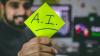 a person at a desk holding a post-it note with 'AI' written on it in thick black pen